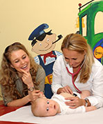 Kinderärztin Dr. med. Claudia Schramm horcht mit einem Stetoskop den Atem eines Babys. Die Mutter des Kindes steht zuversichtlich lächelnd anbei.